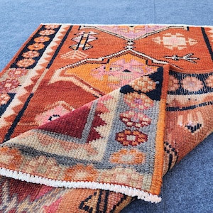 May include: A rectangular woven rug featuring a vibrant orange base with geometric patterns in pink, yellow, and dark blue. The rug has a folded corner revealing a textured underside, a dark blue border with circular motifs, and white fringe edges.