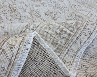 May include: Close-up of a beige and brown patterned rug with a rolled edge. The rug features a detailed design with geometric and floral motifs. The fringe is white, and the rug appears to be made of a woven material.