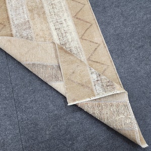 May include: A rectangular runner rug featuring a beige and tan color palette with a repeating zigzag pattern and vertical striped sections. The rug is folded at one corner, revealing the woven texture and the underside of the fabric material.