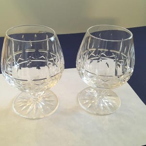 May include: Two clear crystal brandy snifters with intricate cut glass patterns. The glasses have a rounded bowl shape and sit on a short stem with a starburst design. They are placed on a white surface.