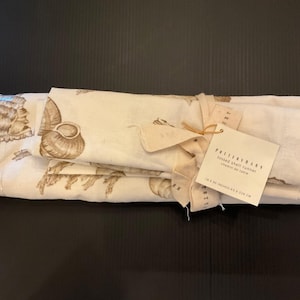 May include: A folded off-white table runner featuring a brown seashell print, tied with a fabric ribbon and a tag labeled Pottery Barn tossed shell runner 18 x 90 inches / 46 x 228 cm. The textile displays various detailed shell illustrations.
