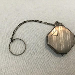 May include: Silver-toned octagonal compact with a chain and ring. The compact has vertical stripes and a triangle design. The chain is attached to a ring. The compact is likely vintage.