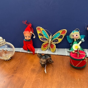 May include: A set of six vintage Christmas ornaments, including a clear glass ornament with a pearl-like top, a red elf ornament, a green angel ornament, a butterfly ornament with iridescent wings, a brown and black dog ornament, and a red and green drum ornament.