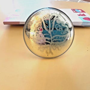 May include: A clear glass paperweight with a silver-colored rim. The paperweight has a design of a tree with a blue background and pink and white accents. The paperweight is on a light brown surface.