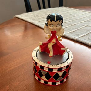May include: A Betty Boop kitchen timer featuring a figure in a red dress with white fur trim standing atop a cylindrical base. The base has a black and white diamond pattern, a silver top, and a numbered dial ranging from 0 to 55 minutes.