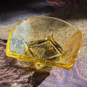 May include: A clear glass bowl with a square base and a fan-shaped top. The bowl has a textured surface and a golden-yellow rim. The bowl is sitting on a purple and brown patterned surface.