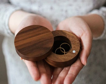 Puede incluir: Una caja de anillos de madera con la tapa abierta, revelando dos anillos de oro en el interior. La caja está hecha de madera oscura y tiene un acabado liso.