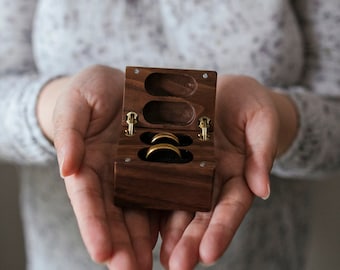 Peut inclure: Une boîte à anneaux en bois avec deux anneaux en or à l'intérieur. La boîte est ouverte et les anneaux sont visibles. La boîte est tenue dans les mains d'une personne.