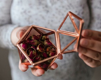 May include: A rose gold geometric glass box with a hinged lid, filled with dried rose buds and gold rings.