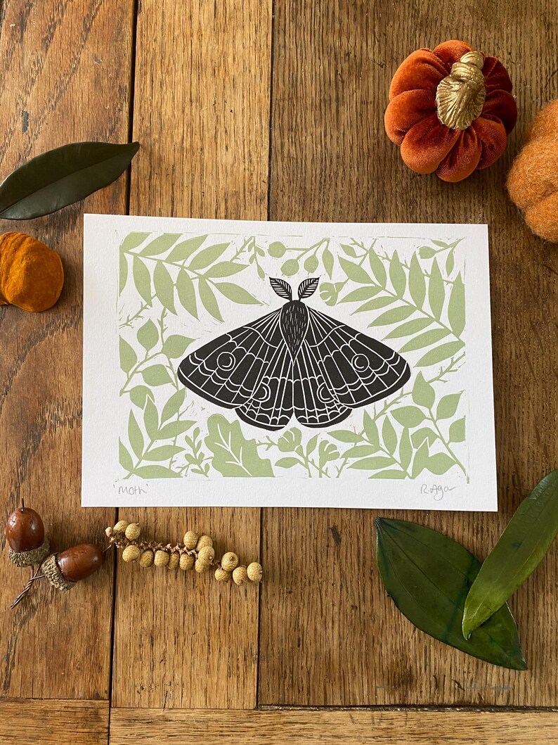 May include: A black and white moth print on a white background with a light green leaf pattern. The print is on a wooden surface with decorative pumpkins, leaves, and acorns. The word "moth" is printed on the bottom left.