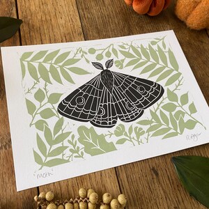 May include: A black and white moth print on white paper, set against a light green leaf pattern. The word "moth" is printed at the bottom left. The print is on a wooden surface with decorative pumpkins and leaves.