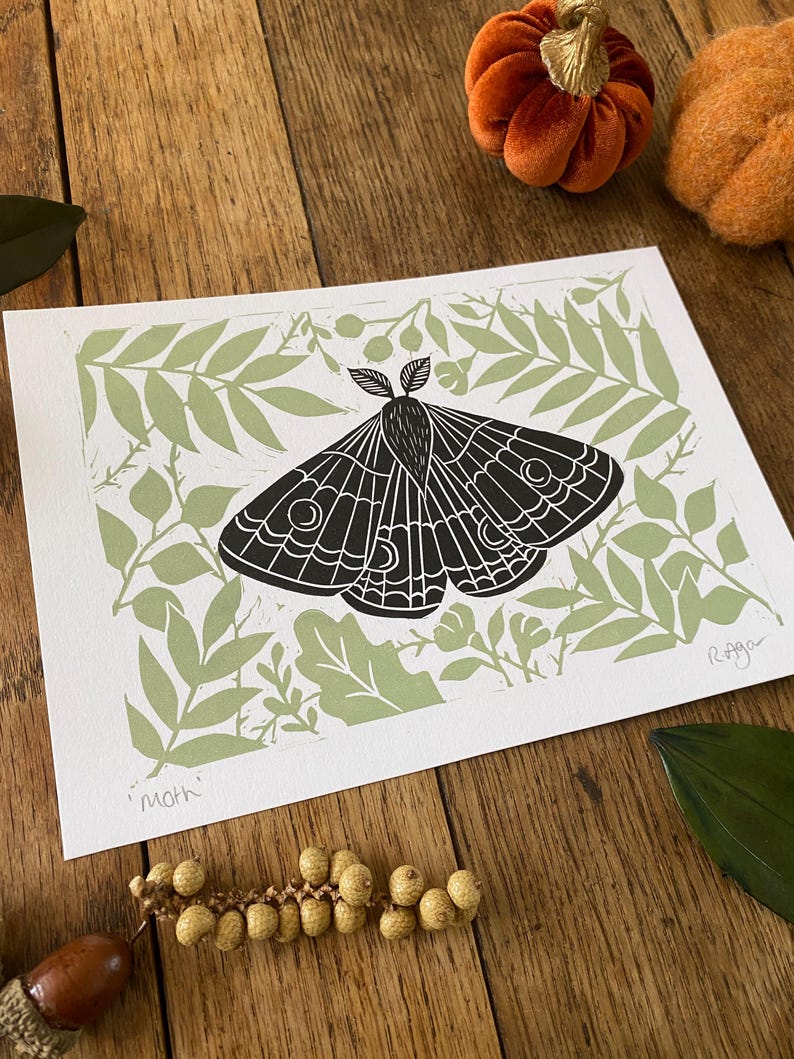 May include: A black and white moth print on white paper, set against a light green leaf pattern. The word "moth" is printed at the bottom left. The print is on a wooden surface with decorative pumpkins and leaves.