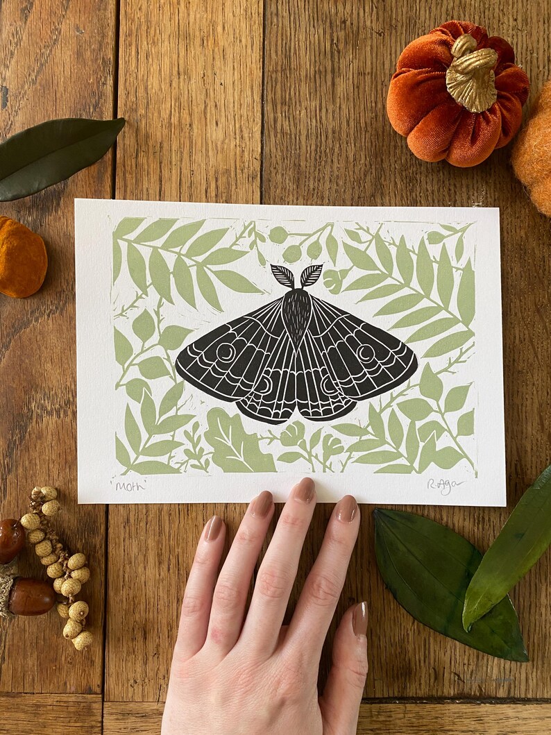 May include: A black and white moth print on white paper, set against a light green botanical background. The word "moth" is printed in the lower left corner. The print is displayed on a wooden surface with decorative pumpkins and leaves.