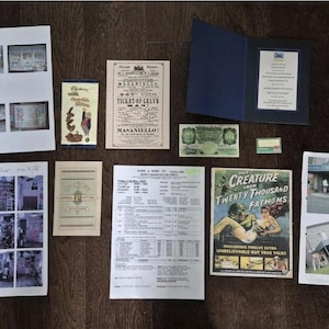 May include: A collection of vintage ephemera, including a Cadbury's chocolate wrapper, a ticket, a movie poster for "The Creature from Twenty Thousand Fathoms", and various documents and photographs, all arranged on a dark wooden surface.