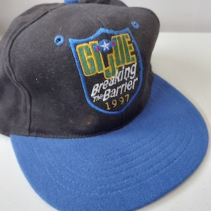 Könnte beinhalten: Eine schwarze und blaue Baseballkappe mit einem gestickten G.I. Joe-Logo auf der Vorderseite. Das Logo ist ein Schild mit einem Stern und den Worten "Breaking the Barrier" und "1997".