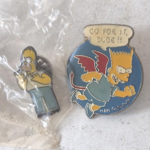 Könnte beinhalten: Zwei Emaille-Anstecknadeln mit Figuren aus den Simpsons. Eine Anstecknadel zeigt Homer Simpson in einem gelben Hemd und blauen Hosen. Die andere Anstecknadel zeigt Bart Simpson als Teufel mit dem Text "GO FOR IT, DUDE !!".