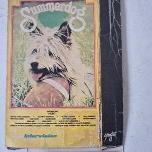 Peut inclure: Un boîtier VHS vintage pour le film Summerdog, illustré par un chien hirsute avec une balle. La couverture jaune porte le texte Summerdog is kid's best friend et Summerdog is family fare, ainsi que les crédits de production et du casting.