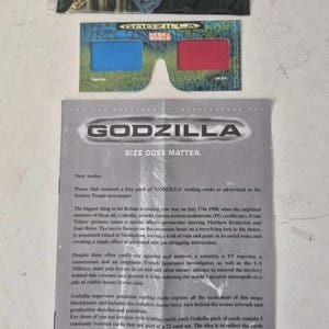 Può includere: Immagine promozionale del film Godzilla, con un pacchetto di figurine, occhiali 3D e una lettera. Il pacchetto di figurine presenta Godzilla. Gli occhiali 3D hanno lenti rosse e blu. La lettera recita "SIZE DOES MATTER."