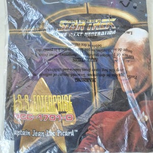 May include: A Star Trek: The Next Generation t-shirt in a plastic bag. The shirt features Captain Jean-Luc Picard and the U.S.S. Enterprise NCC-1701-D. The shirt is blue with a graphic design.