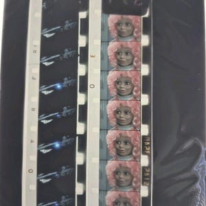 Puede incluir: Una tira de película con múltiples fotogramas. Una tira muestra una nave espacial en un espacio oscuro con un brillo azul. La otra tira muestra una muñeca con cabello rizado rosa y un atuendo rosa. La tira de película está en un marco negro.