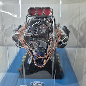 Może przedstawiać: Szczegółowy model silnika Ford Dragster, zamknięty w przezroczystym plastiku. Silnik posiada chromowane elementy, czerwone akcenty i skomplikowane okablowanie. Podstawa jest niebieska z logo Ford Dragster engine.