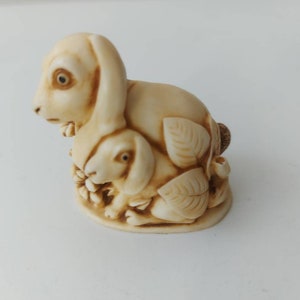 May include: A cream-colored ceramic figurine of a mother rabbit and her baby rabbit. The mother rabbit is sitting upright with her ears perked up, while the baby rabbit is nestled in her side. Both rabbits have brown eyes and are surrounded by leaves.