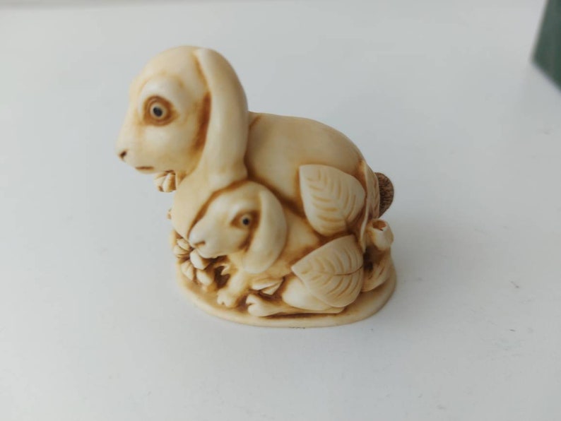 May include: A cream-colored ceramic figurine of a mother rabbit and her baby rabbit. The mother rabbit is sitting upright with her ears perked up, while the baby rabbit is nestled in her side. Both rabbits have brown eyes and are surrounded by leaves.