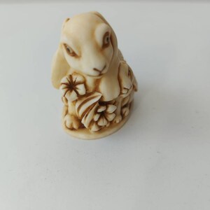 May include: A small, cream-colored ceramic figurine of a rabbit holding flowers. The rabbit has brown eyes and a brown nose.