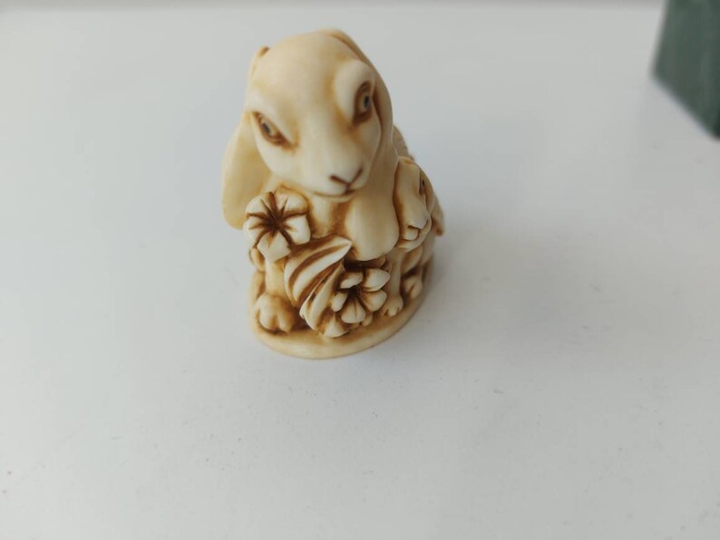May include: A small, cream-colored ceramic figurine of a rabbit holding flowers. The rabbit has brown eyes and a brown nose.