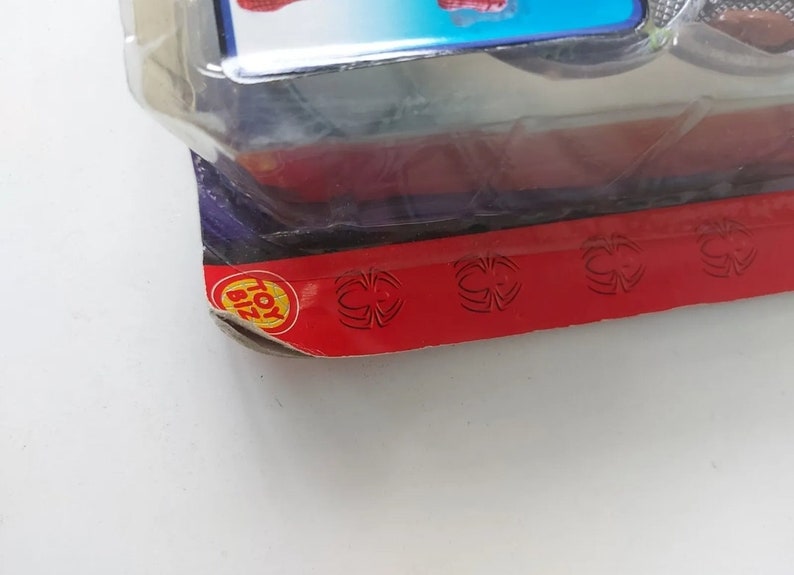 Pode incluir: Close-up de um logotipo Toy Biz vermelho e amarelo em um fundo vermelho com tr&ecirc;s desenhos de teia de aranha preta.