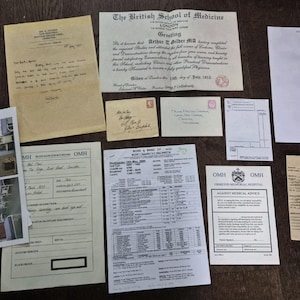 May include: A collection of vintage documents, including letters, certificates, and medical records, are arranged on a dark wooden surface. The documents vary in size and color, with handwritten text and printed elements. Some feature stamps and seals.