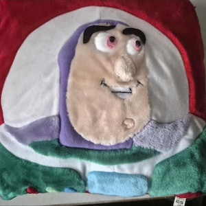 May include: A plush pillow featuring the face of Buzz Lightyear from Toy Story. The pillow has a red, white, green, purple, and light blue color scheme. The character's face is in the center, with a smiling expression.
