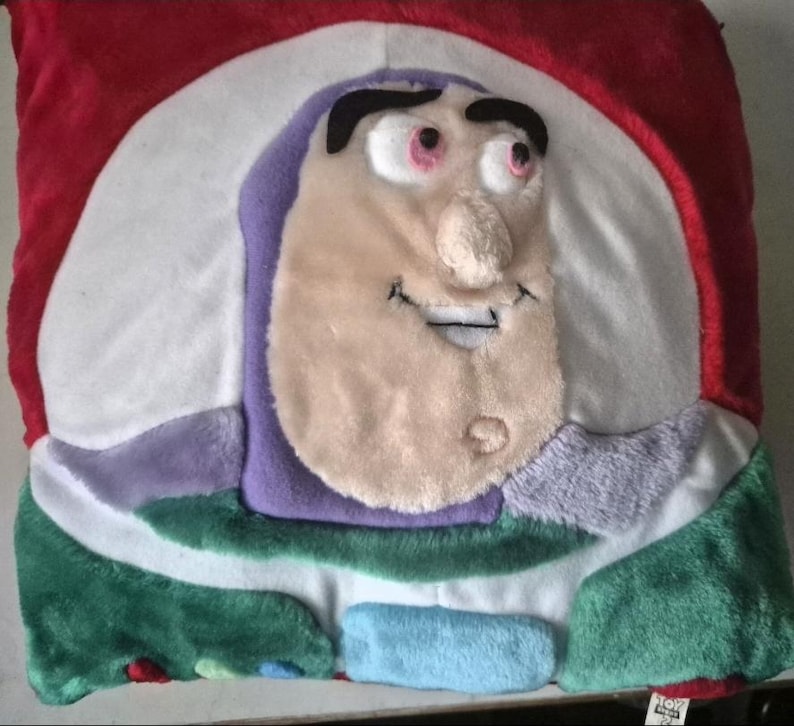 May include: A plush pillow featuring the face of Buzz Lightyear from Toy Story. The pillow has a red, white, green, purple, and light blue color scheme. The character's face is in the center, with a smiling expression.