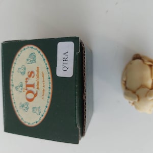 May include: A small, cream-colored ceramic figurine of a pig in a curled-up position. The figurine is in a box with the text "The QT's Animal Collection A Noble Art Product" and "QTRA" on it.
