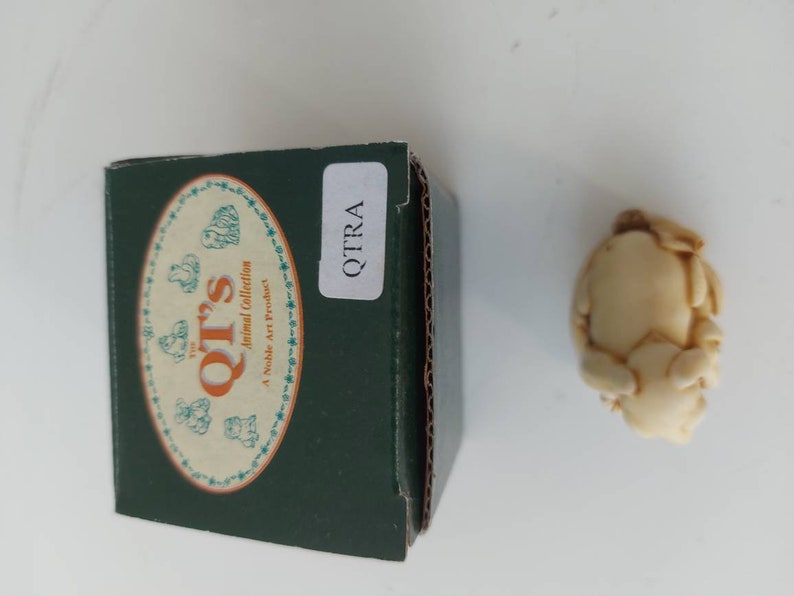 May include: A small, cream-colored ceramic figurine of a pig in a curled-up position. The figurine is in a box with the text "The QT's Animal Collection A Noble Art Product" and "QTRA" on it.