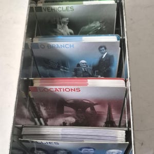 以下が含まれることがあります： 金属製のカードホルダーに、カードゲームのデッキが詰まっています。「Villains」、「Vehicles」、「IO Branch」、「Locations」、「Allies」などのカテゴリーでラベル付けされたカード。カードには画像とテキストが表示されており、テーマ別のカードゲームを示唆しています。
