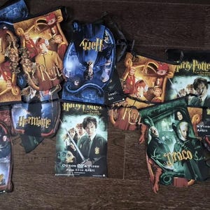 Puede incluir: Una colección de artículos temáticos de las películas de Harry Potter, que incluyen estuches de DVD y materiales promocionales. Los artículos presentan nombres de personajes y títulos de películas, con colores vibrantes e imágenes del elenco. Los artículos están esparcidos sobre una superficie de madera.