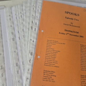 May include: A stack of white plastic sheet protectors containing a shooting script for the television show Spooks, Episode Two, by David Wolstencroft, dated Friday 2nd November 2001, printed on an orange cover page with black text listing production staff.