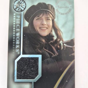 May include: A trading card featuring a smiling woman wearing a black beret and coat. The card has the text "PIECE WORKS" and a textured black patch. The card has a light blue and silver color scheme.