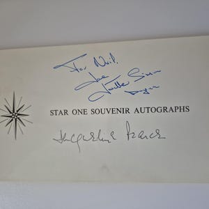 Op de afbeelding: Een vintage souvenir autograafkaart met blauwe inkt handtekeningen en de tekst "STAR ONE SOUVENIR AUTOGRAPHS". Een zwart-witte starburst grafiek staat links. De kaart heeft een crèmekleurige achtergrond.