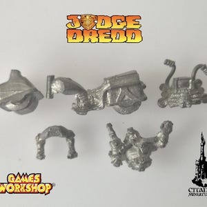 May include: Miniature metal Judge Dredd model kit pieces. The image shows various components, including a helmet, motorcycle, and other parts. The text "Judge Dredd" and "Games Workshop" are visible.
