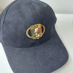 Könnte beinhalten: Eine schwarze Baseballkappe mit einem goldfarbenen Aufnäher, der ein Schild mit dem Text "35 Years" und "G.O.P.E." zeigt. Das Schild ist außerdem in Rot, Weiß und Blau gehalten. Der Text "1964 - 1999" befindet sich unterhalb des Schildes.