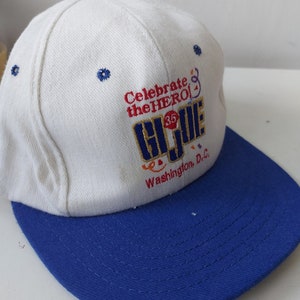 Könnte beinhalten: Weiße Baseballkappe mit blauem Schirm. Auf der Vorderseite der Kappe befindet sich ein gesticktes Logo mit der Aufschrift "Celebrate the HERO! G.I. JOE 35 Washington, D.C."