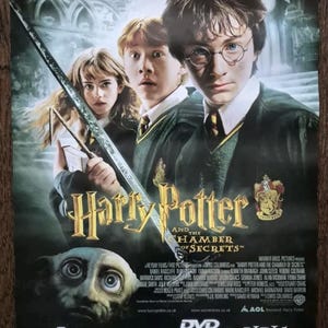 Puede incluir: Un póster de película para "Harry Potter y la Cámara Secreta", con Daniel Radcliffe, Rupert Grint y Emma Watson como Harry Potter, Ron Weasley y Hermione Granger, respectivamente. El póster también presenta el escudo de Hogwarts y el texto "Disponible en DVD y VHS a partir del 11 de abril".