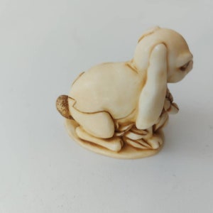 May include: A small, cream-colored ceramic figurine of a rabbit sitting on its haunches with its ears folded back. The rabbit is holding a bunch of small, brown leaves in its paws.