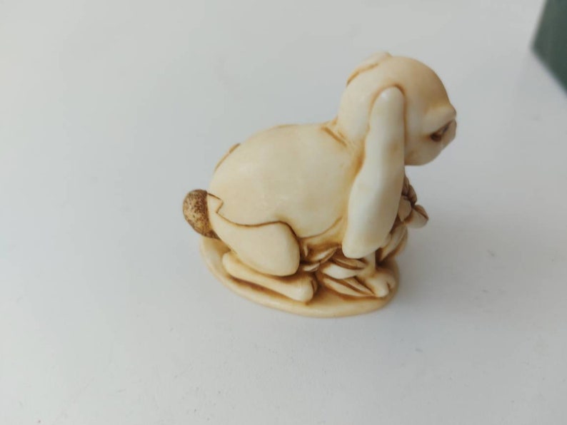 May include: A small, cream-colored ceramic figurine of a rabbit sitting on its haunches with its ears folded back. The rabbit is holding a bunch of small, brown leaves in its paws.