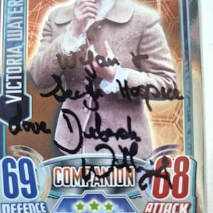 May include: A collectible trading card featuring Victoria Waterfield, with a score of 69 for Defense and 68 for Attack. The card has a photo of a woman in a brown jacket and is signed in black ink.