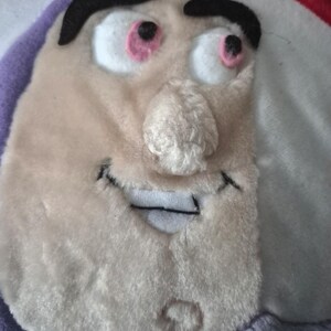 May include: A plush toy featuring the face of a cartoon character with a light beige complexion, black eyebrows, and pink-rimmed eyes. The toy has a stitched smile and a nose with a textured appearance. The background includes purple, white, and red fabric.