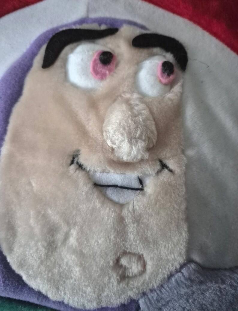 May include: A plush toy featuring the face of a cartoon character with a light beige complexion, black eyebrows, and pink-rimmed eyes. The toy has a stitched smile and a nose with a textured appearance. The background includes purple, white, and red fabric.