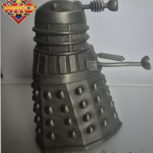 Könnte beinhalten: Graue metallische Dalek-Figur aus der Doctor Who-Serie. Die Figur hat einen zylindrischen Körper mit zahlreichen runden Nieten, einen Netzmittelteil und zwei armartige Anhänge. Das Doctor Who-Logo ist sichtbar.